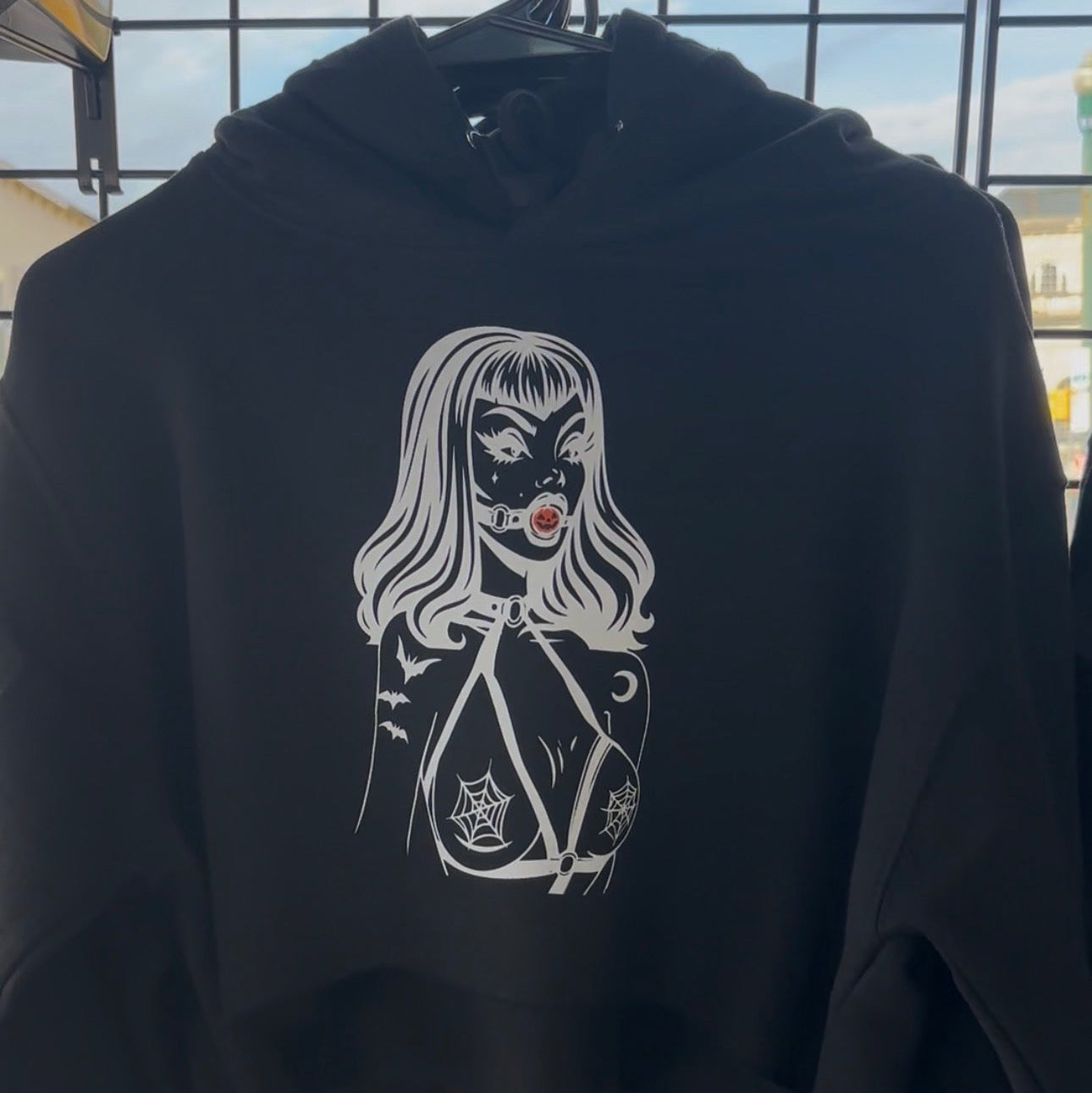 A black cropped hoodie hanging on a black hanger, featuring a white graphic print of a gothic-style woman with a skull and crossbones on the front. The hoodie is photographed outdoors against a black wire grid fence with sky visible in the background.